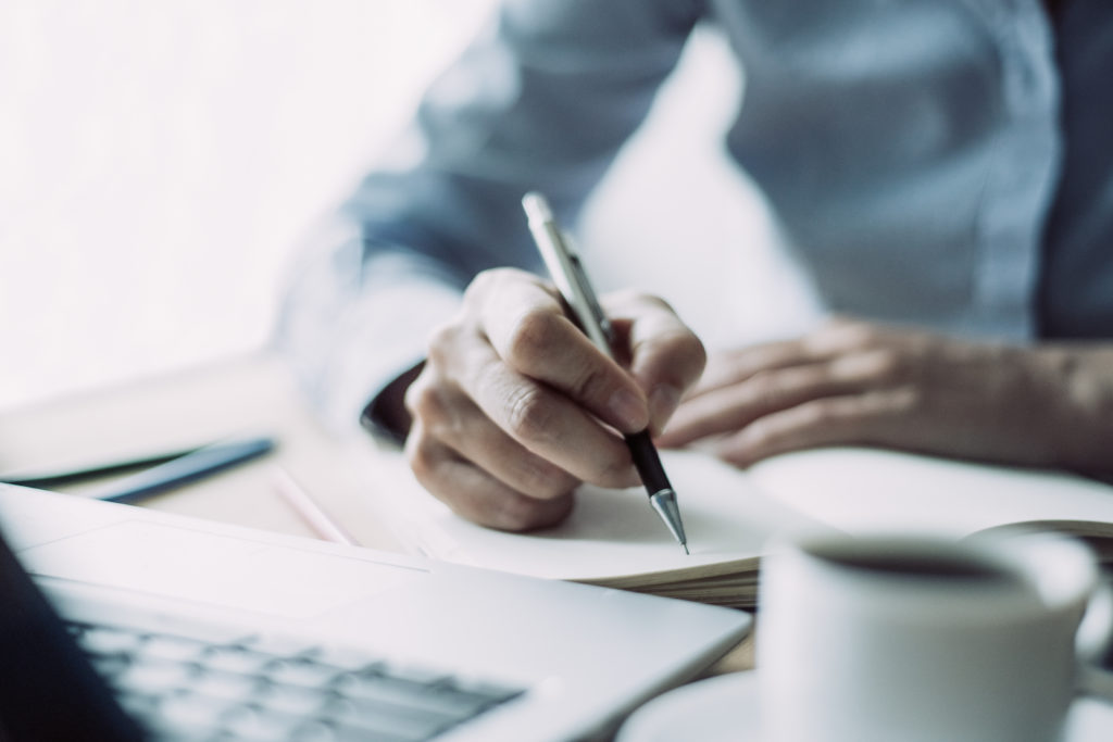 Cropped view of woman writing in notebook and sitting at desk with laptop computer